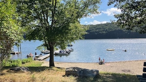 A lakeside view with boats docked, a sandy beach, and a large tree providing shade. - New Build, Private Beach, Central Air, Big Yard, Firepit, Wine Bar (Narragansett)