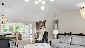 A spacious living room with a grey sofa, a marble coffee table, and a dining area with a large table and chairs. - New Build, Private Beach, Central Air, Big Yard, Firepit, Wine Bar (Narragansett)