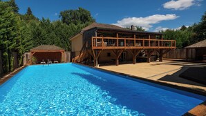 Ein Schwimmbecken mit Holzterrasse, einer überdachten Sitzgruppe und einem Gebäude mit geneigtem Dach.
