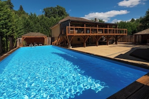 A swimming pool with a wooden deck, a covered seating area, and a building with a sloped roof.