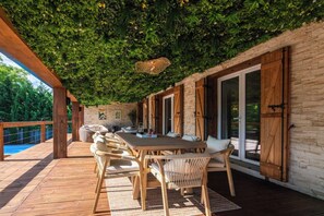 Eine Außenpatio mit Holztisch und -stühlen, einer Pergola mit Kletterpflanzen und Blick auf ein Schwimmbad.