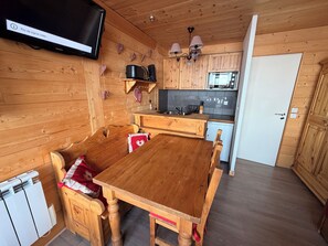 Un interior de madera con una mesa de comedor, sillas, un televisor en la pared, una zona de cocina con armarios y electrodomésticos, y una puerta.