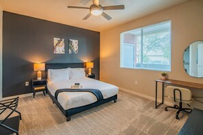 A hotel room with a bed, bedside lamps, a desk, a chair, a ceiling fan, and artwork on the wall.