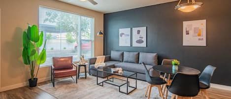 A modern living room with a sofa, coffee table, and chairs, a dining table with chairs, a ceiling fan, and artwork on the walls.