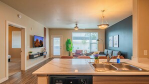 A modern kitchen with a dishwasher, a dining area with a table and chairs, a living room with a sofa, and a large window with blinds. - Tempe DT Escape | Pool, Gym, W/D, Parking Sleeps 4 (Tempe)