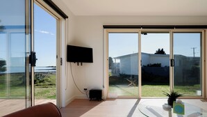 A modern living room with a glass table, a flat-screen TV mounted on the wall, and a sliding glass door leading to a backyard.