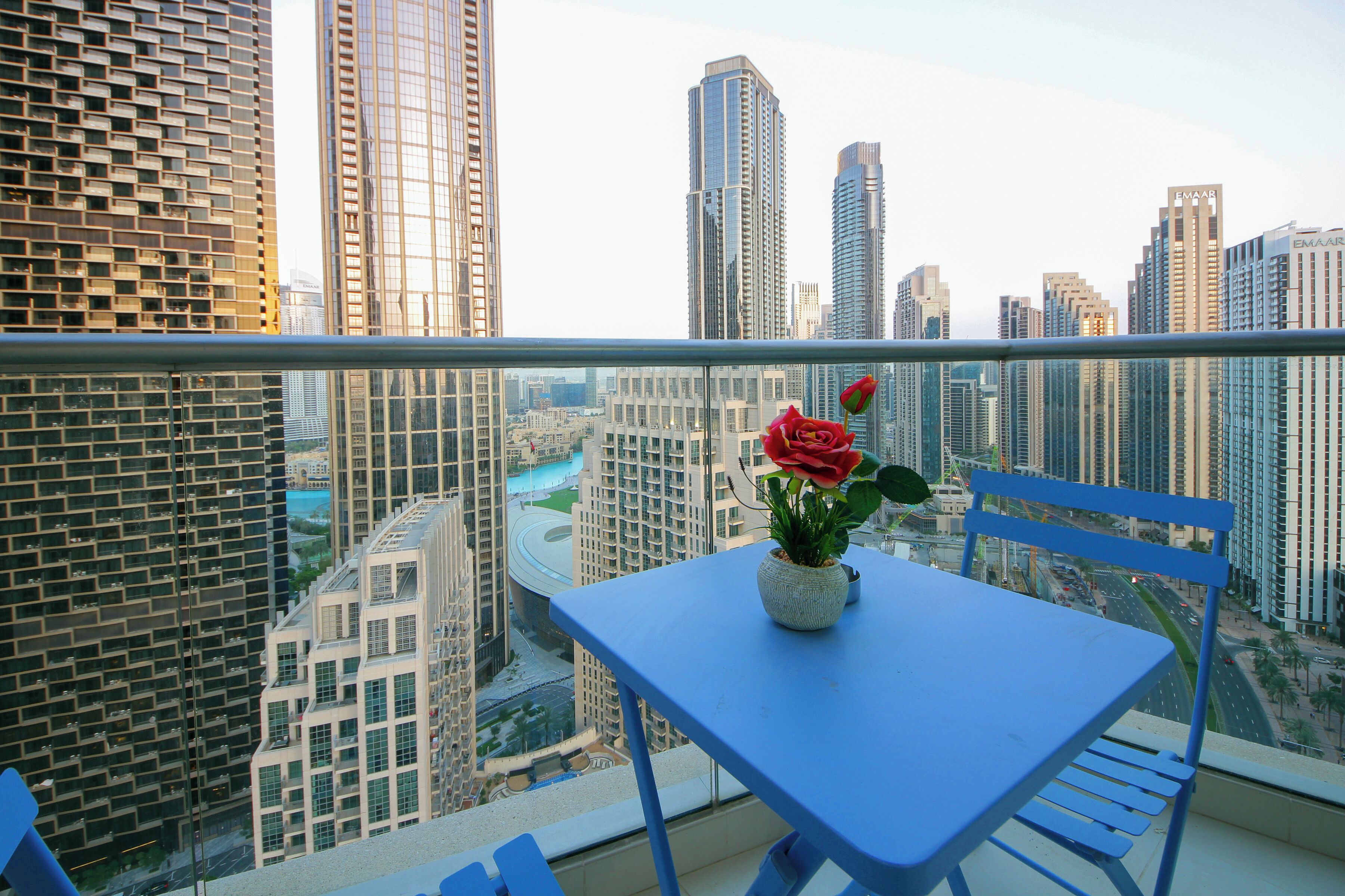 A balcony with a table and chair, a potted plant with a red rose, and a cityscape view.