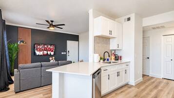 A modern kitchen with white cabinetry, a built-in dishwasher, and a gray sofa in the living area.