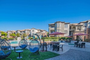 A poolside area with hanging chairs, tables, and umbrellas, surrounded by multi-story residential buildings. - 3Min To Stadium w/ Balcony Pool Gym Parking W/D (Glendale)