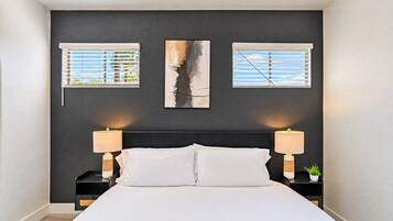 Une chambre à coucher avec un lit, deux tables de chevet avec des lampes et trois fenêtres équipées de stores.