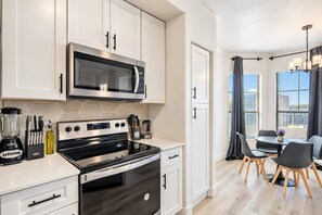 A modern kitchen with white cabinetry, a stainless steel microwave, and a stove. There is a dining area with a table and chairs, and a view of buildings through large windows. - 3Min To Stadium w/ Balcony Pool Gym Parking W/D (Glendale)