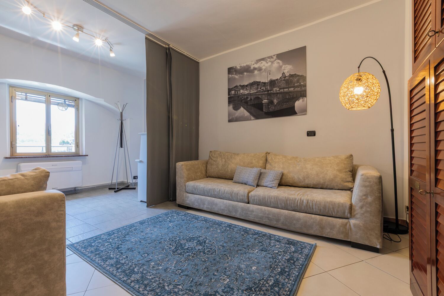 A modern living room with a beige sofa, a blue rug, and a cityscape painting on the wall.