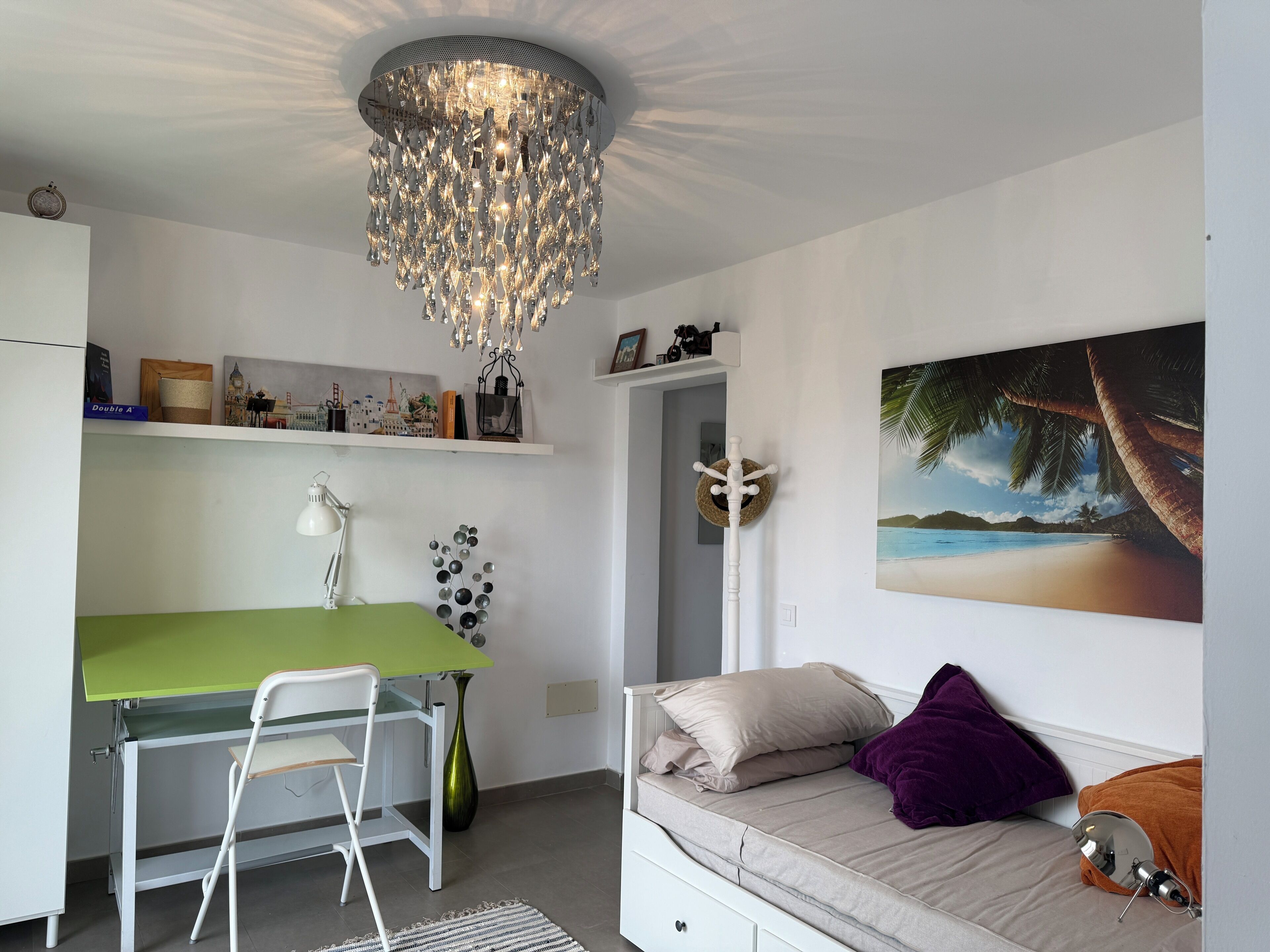 A bedroom with a bed, a desk with a chair, a green table, a chandelier, a painting of a beach, and a door with a coat rack.