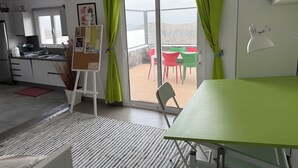 Une cuisine moderne avec des meubles blancs, un coin repas avec des chaises vertes et une vue sur l’extérieur grâce à de grandes fenêtres.