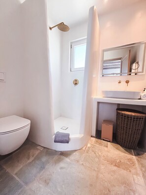 A bathroom with a white toilet, a shower with a gold showerhead, a sink with a mirror above it, and a wicker basket on the floor. - Elysian Villas Ios (Ios)