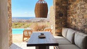 Un patio con una mesa de piedra, una lámpara colgante de mimbre, un sofá con cojines y vistas a una piscina y al paisaje.