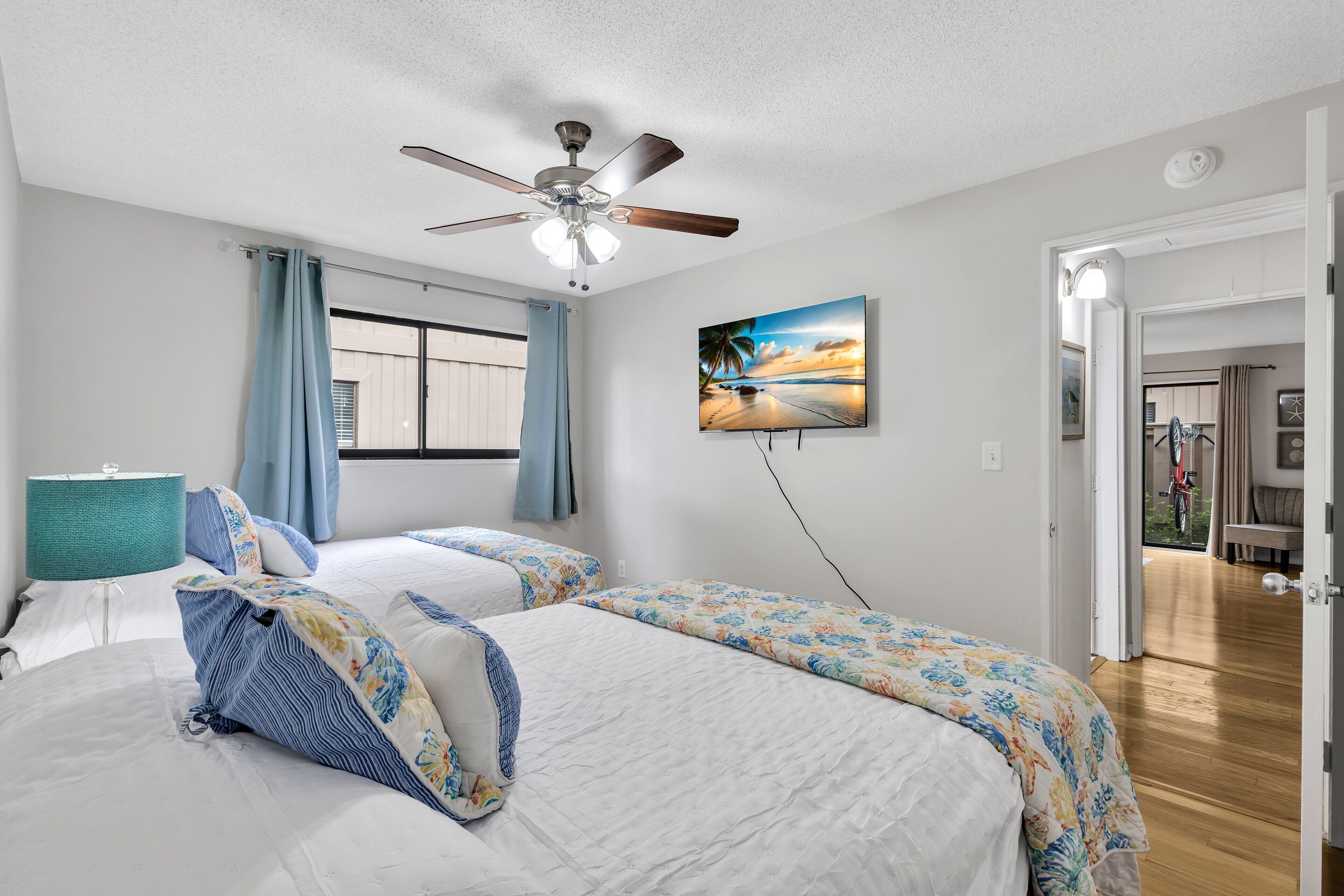 Une chambre avec deux lits, un ventilateur de plafond, une fenêtre avec des rideaux et un tableau représentant une scène de plage.