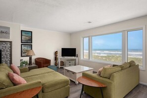 Sala de estar com uma grande janela vista para a praia, televisão e um espaço de estar aconchegante.