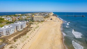 An aerial view of a coastal area with a beach, a pier, and several buildings including a hotel.
