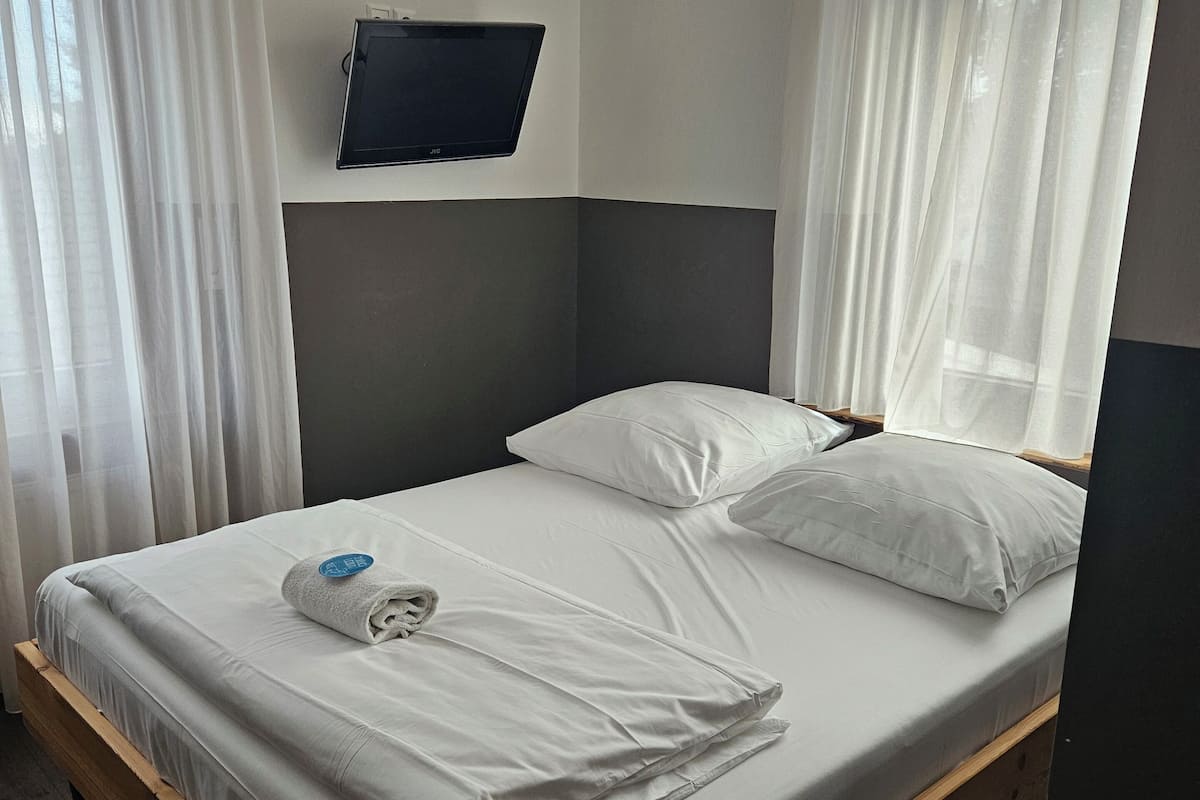 A neatly made bed with white linens and pillows, a flat-screen TV mounted on the wall, and sheer curtains on the windows.