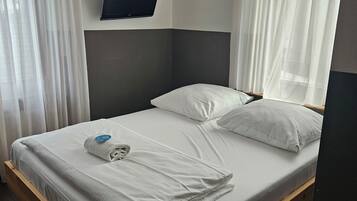 Una cama bien hecha con sábanas y almohadas blancas, un televisor de pantalla plana montado en la pared y cortinas traslúcidas en las ventanas.