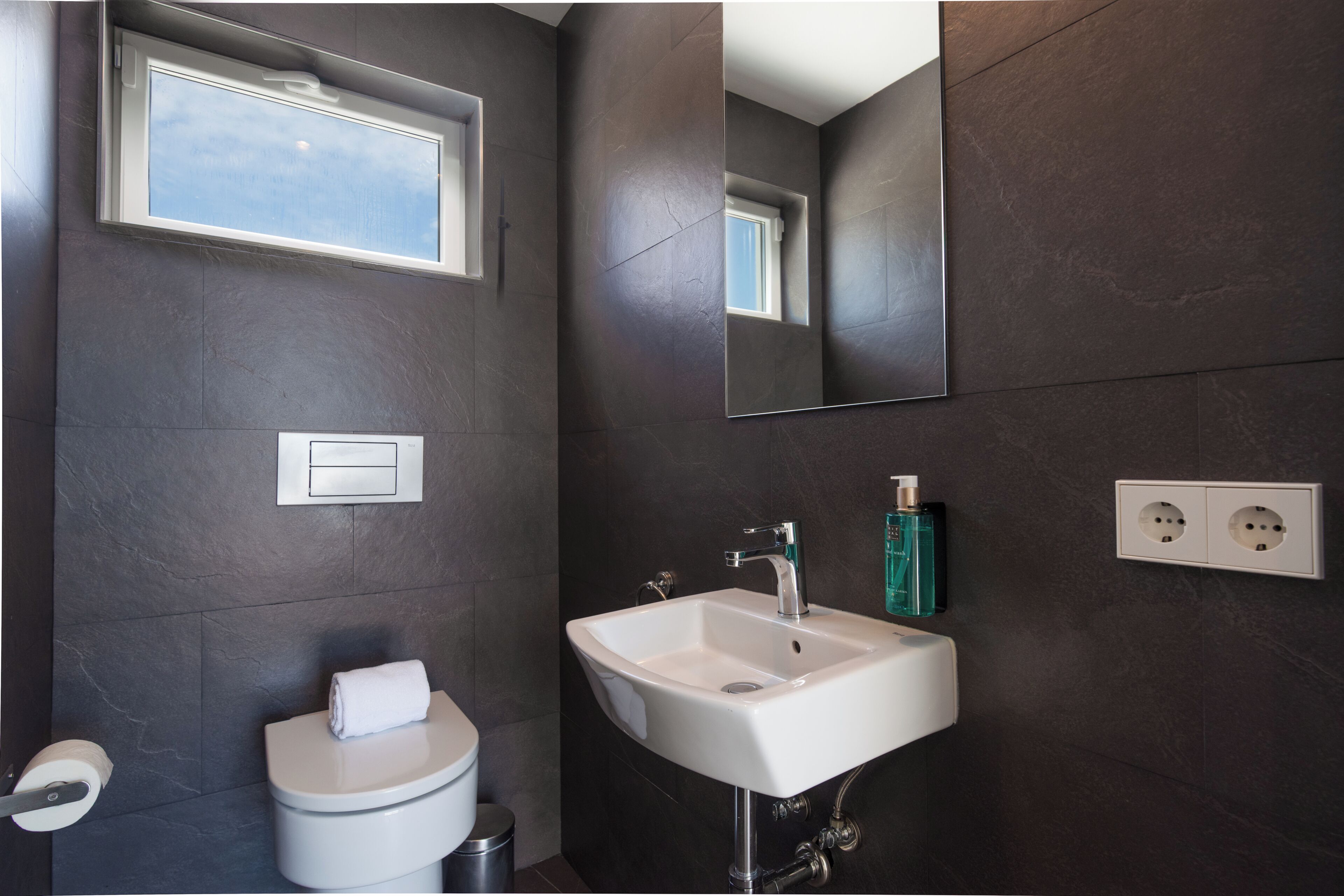 Ein Badezimmer mit einem weißen Waschbecken, einem Spiegel, einem Fenster und einem Handtuch auf der Toilette.