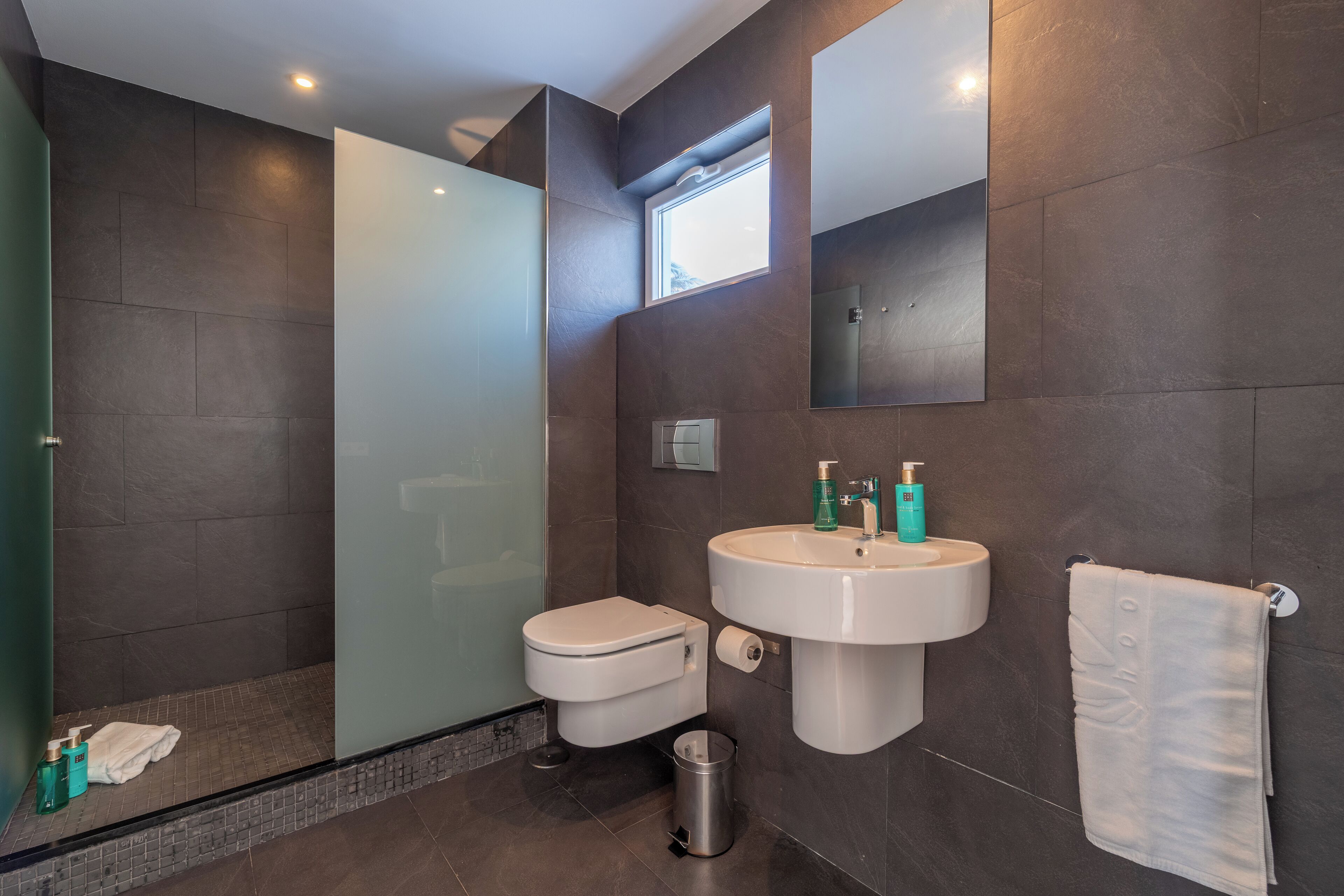 Ein modernes Badezimmer mit einer Duschkabine aus Glas, einer weißen Toilette, einem Waschbecken mit Spiegel darüber und einem Handtuch an einem Halter.
