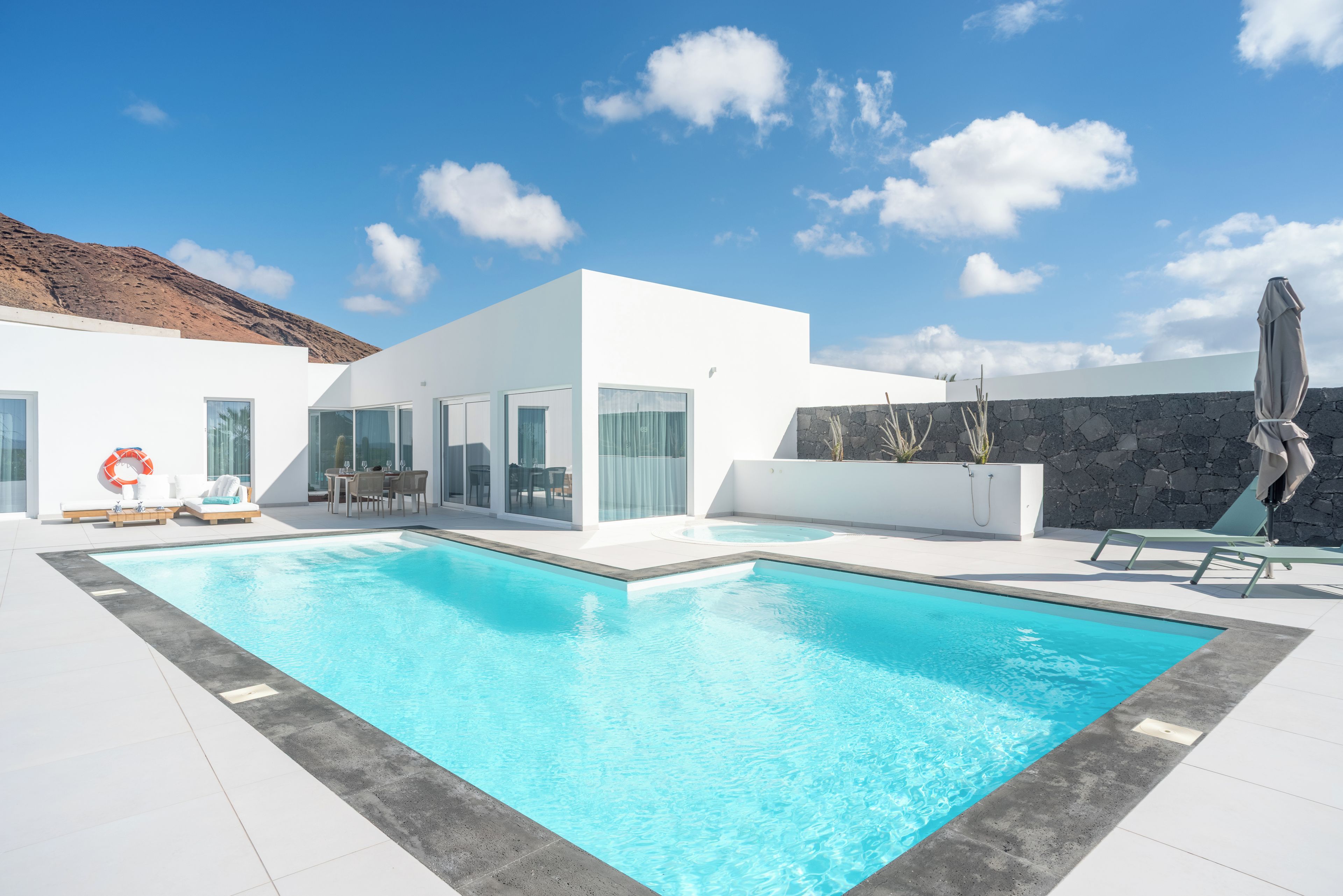 A modern pool area with a clear blue pool, white walls, and a stone wall. There are lounge chairs and a table with umbrellas.