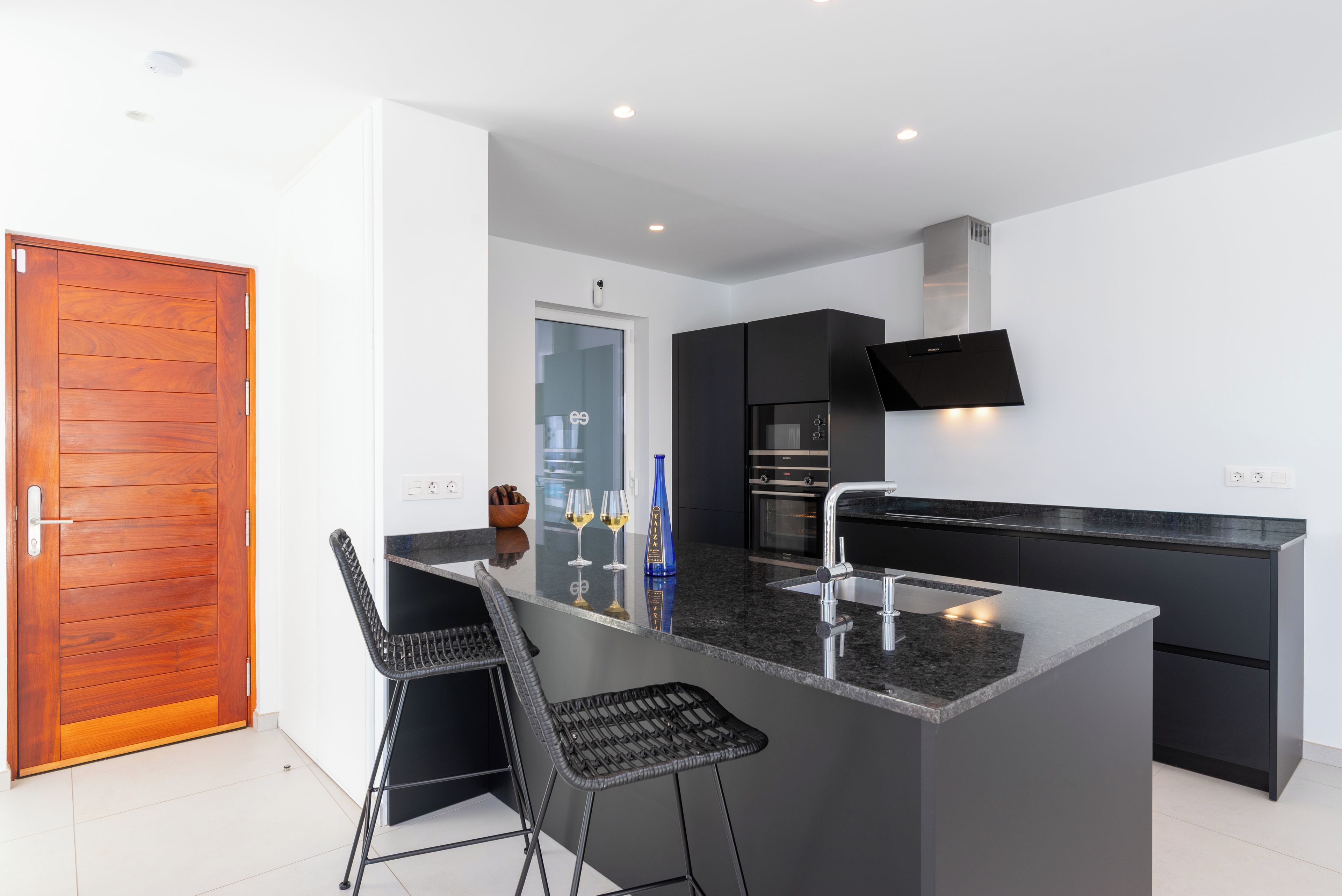 Een moderne keuken met een centraal kookeiland, zwarte barkrukken, een houten deur en een flatscreen-televisie boven het eiland.