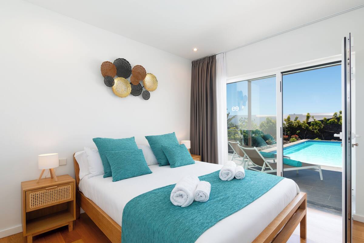 Une chambre d’hôtel moderne avec un grand lit, une literie de couleur turquoise, un élément décoratif mural, une table de chevet et une vue sur une piscine extérieure.