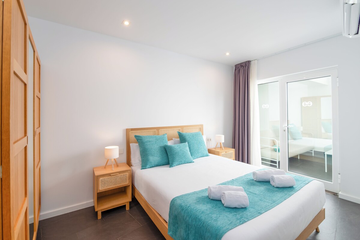 A hotel room with a large bed, wooden headboard, teal bedding, and a balcony with a glass door.