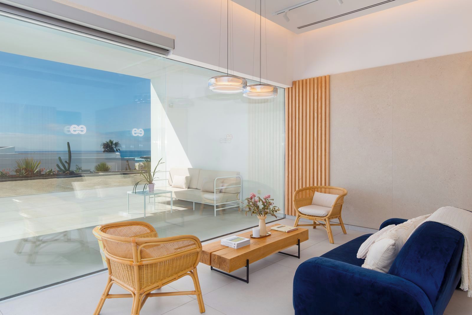 A modern living room with a glass wall, a blue sofa, wicker chairs, a wooden coffee table, and a white couch.