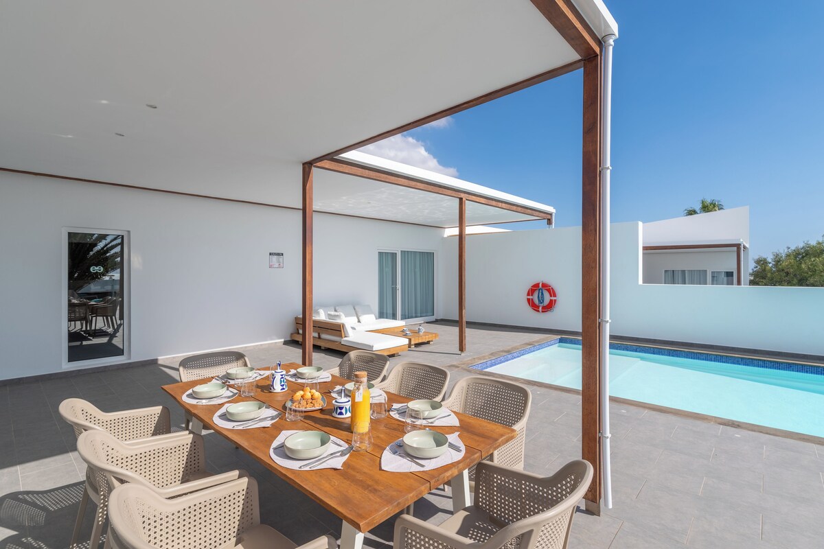 Un área de comedor exterior moderna con piscina, paredes blancas y cielo despejado.