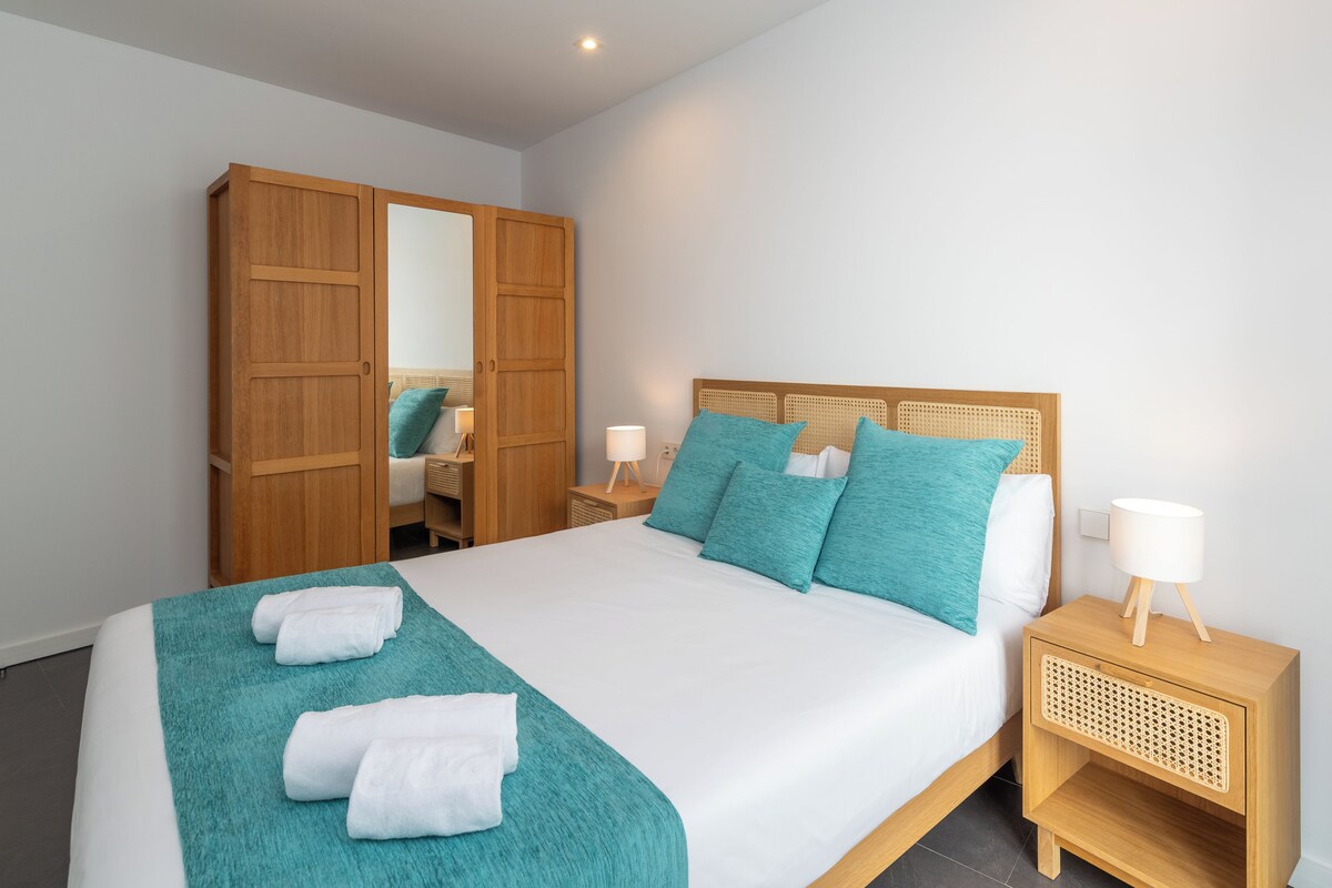 A neatly made bed with a teal blanket and white towels, a wooden wardrobe, and a bedside table with a lamp.