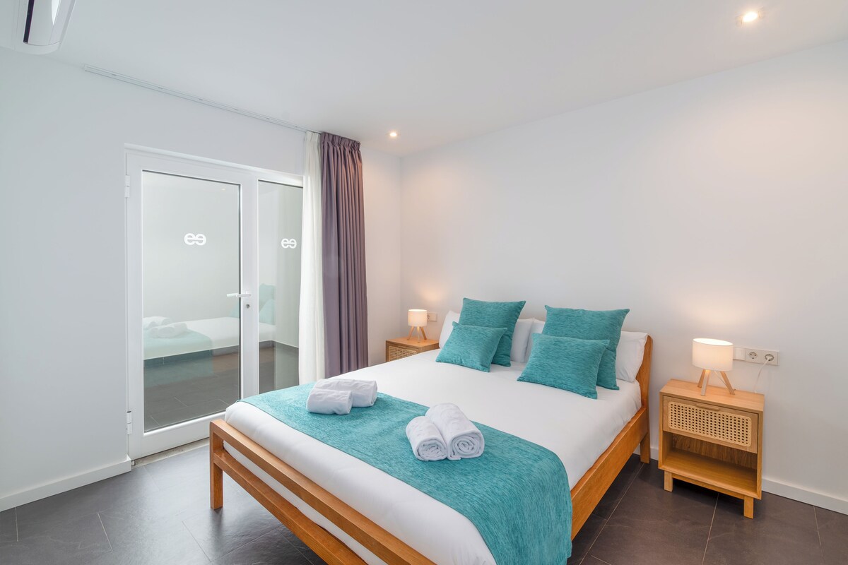 Una camera d'hotel moderna con un letto grande, biancheria da letto verde acqua, una panca in legno, un comodino con una lampada e una porta a vetri scorrevoli.