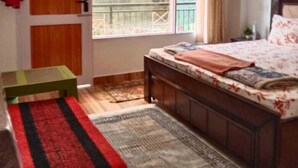 A bedroom with a bed, a small table, a bench, a window with curtains, and a view of trees outside. - Bushahri Homestay (Narkanda)