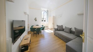 A compact living space with a sofa, a dining table with chairs, a TV mounted on the wall, and a window providing natural light. - Lilla Home (Roma)