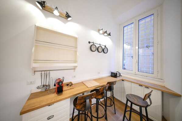 A compact kitchen with a wooden countertop, bar stools, a built-in shelf, and a window with blinds. - Lilla Home (Roma)