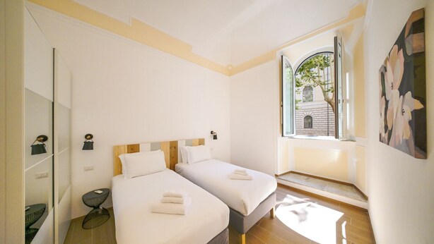 A hotel room with two beds, a window with a view of a building, and a small table with a chair. - Lilla Home (Roma)