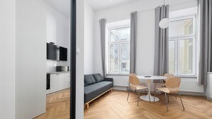 Ein modernes Wohnzimmer mit einer Couch, einem Esstisch mit Stühlen, einem an der Wand montierten Flachbildfernseher und großen Fenstern mit grauen Vorhängen.