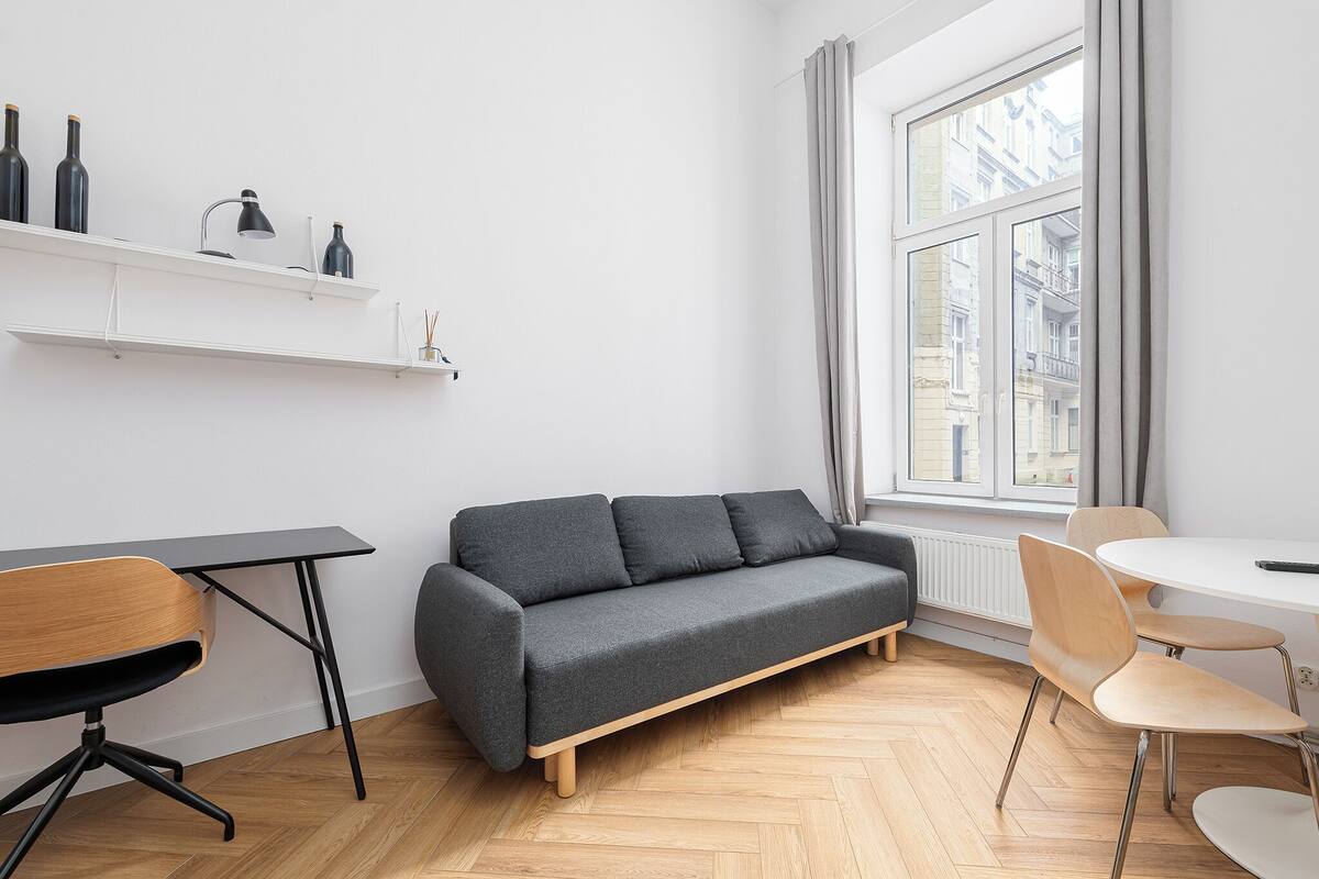 A modern living space with a grey sofa, wooden chairs, a small table, and a white shelf with bottles.