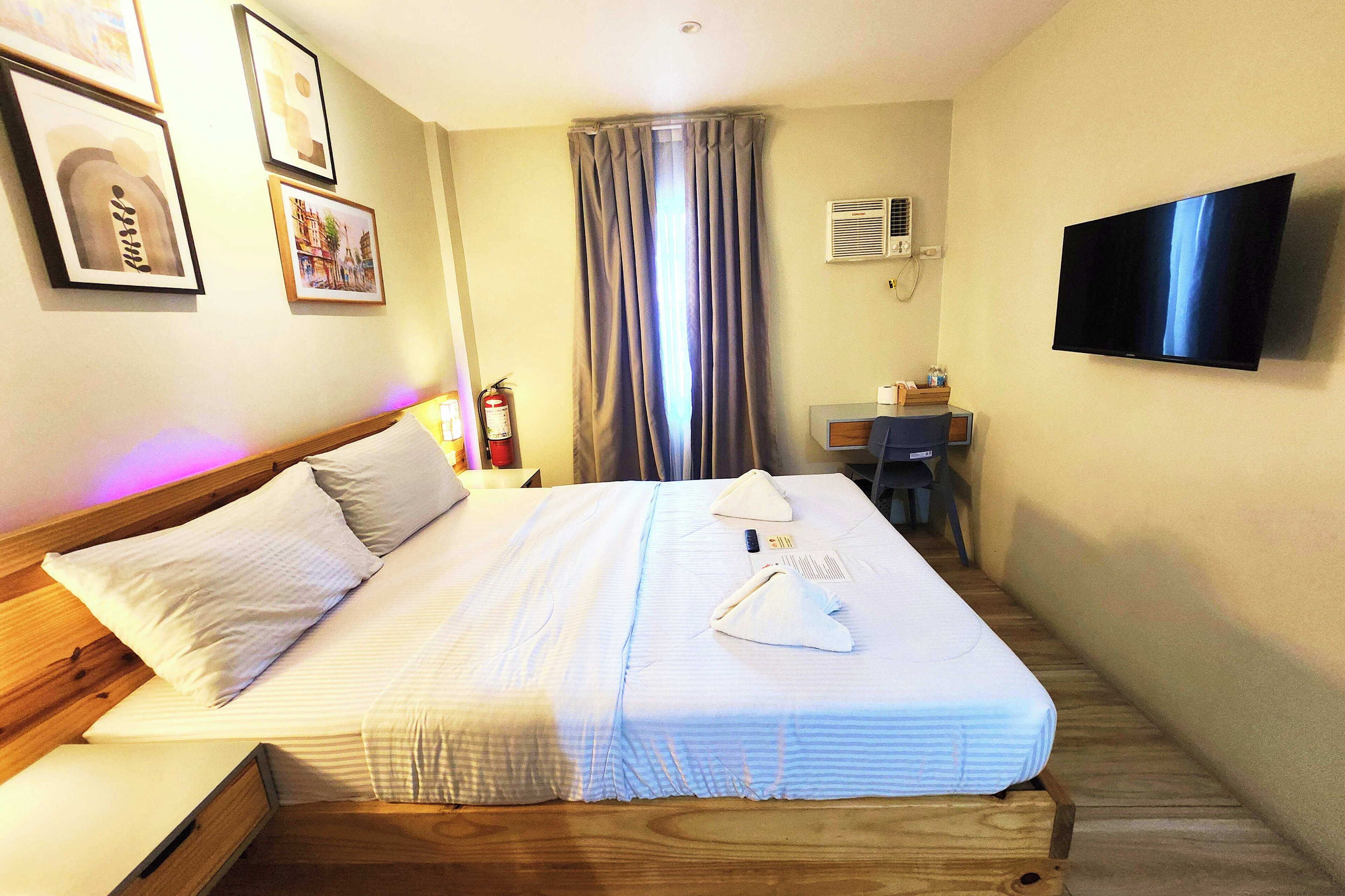 A hotel room with a neatly made bed, a flat-screen TV mounted on the wall, and a desk with a chair.