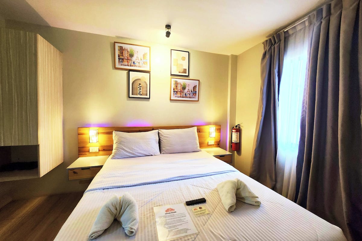 A hotel room with a bed, bedside tables, wall-mounted lamps, framed pictures, a fire extinguisher, and a window with curtains.
