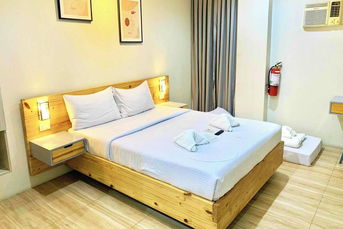 A neatly made bed with white linens, a wooden headboard, bedside tables with lamps, a fire extinguisher, and a small bedside table with a phone and a folded towel.