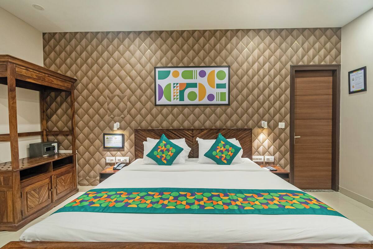 Habitación de hotel con una cama grande, cabecera de madera, panel de pared acolchado, una obra de arte abstracta enmarcada, armario de madera, mesitas de noche con lámparas y una puerta.