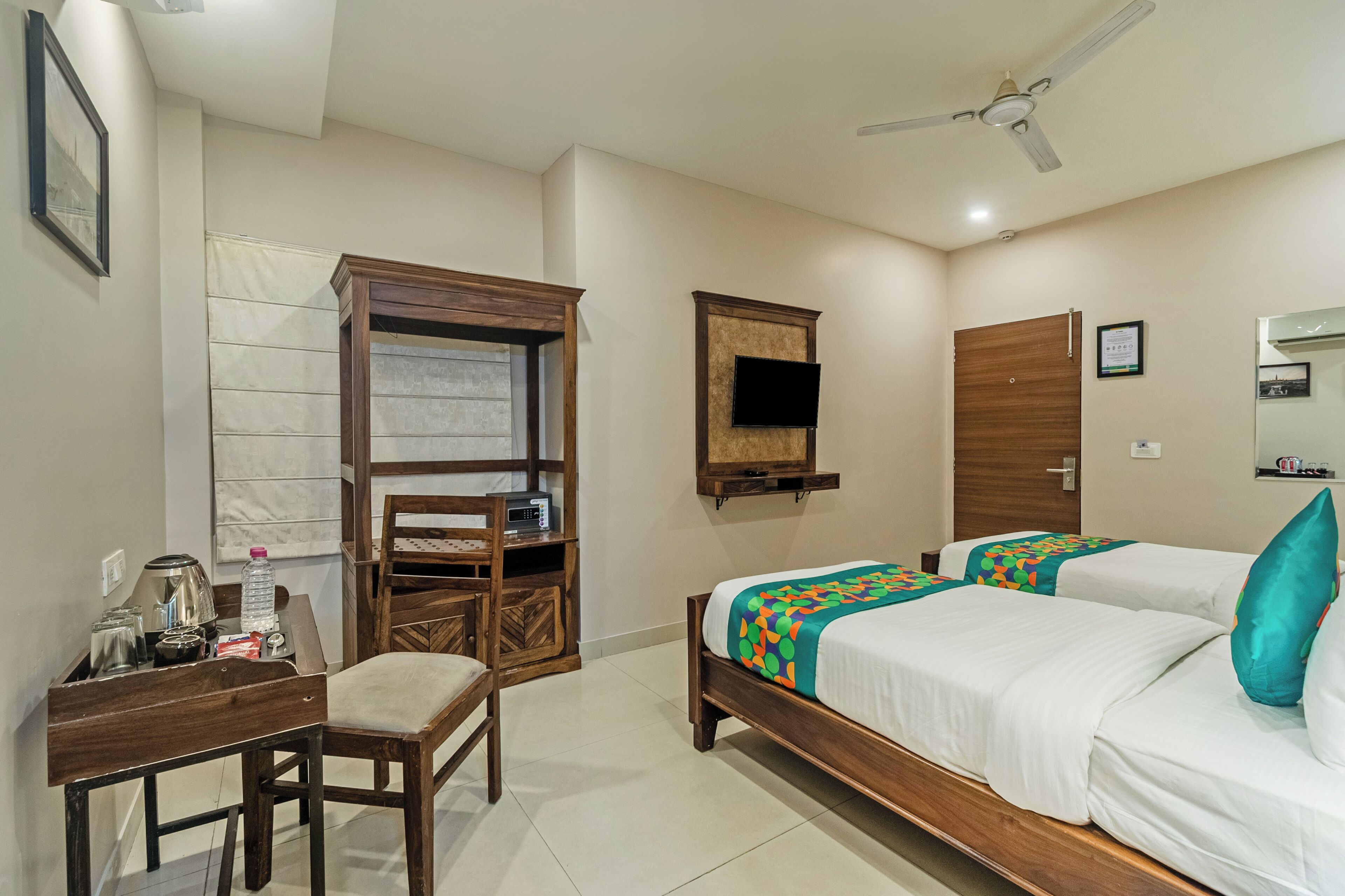 A hotel room with two beds, a desk with a chair, a wardrobe, a television mounted on the wall, and a ceiling fan.
