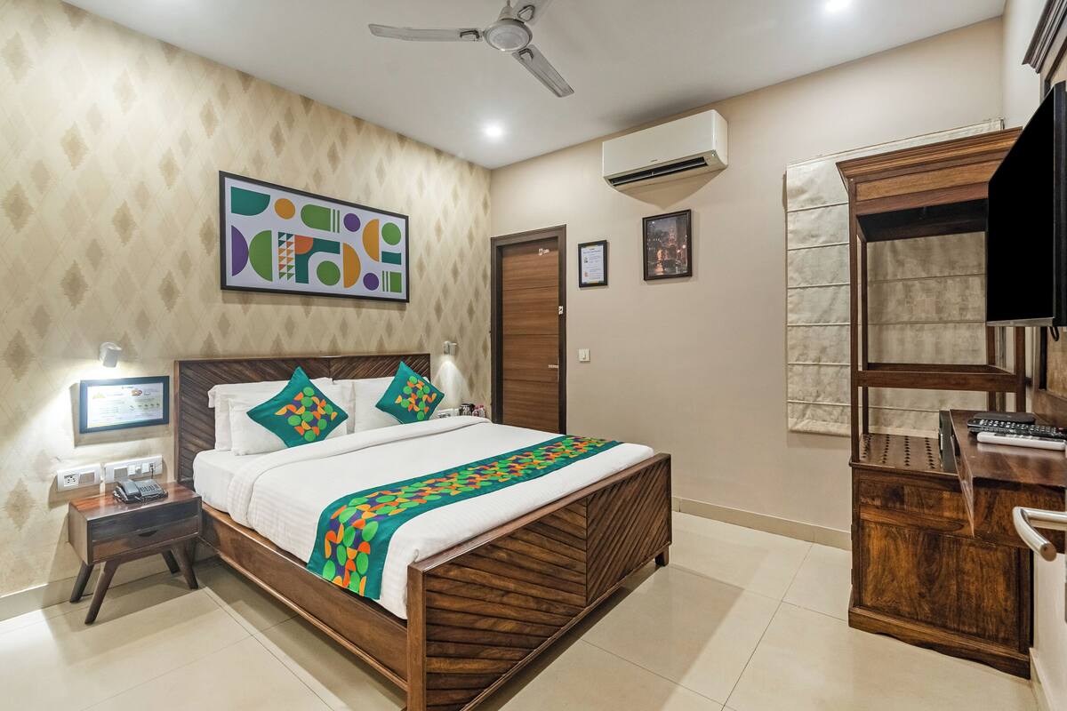 Habitación de hotel con cama de madera, un escritorio con silla, ventilador de techo, unidad de aire acondicionado y un cuadro en la pared.