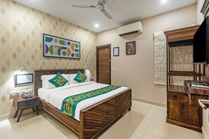 Habitación de hotel con cama de madera, un escritorio con silla, ventilador de techo, unidad de aire acondicionado y un cuadro en la pared.