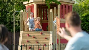 A wooden play structure with climbing holds and a slide, surrounded by a metal fence. - Casita De Cuervo at Country Park (Lymington)