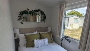 A bedroom with a bed, a bedside lamp, a window with curtains, and a view of outside buildings. - Casita De Cuervo at Country Park (Lymington)